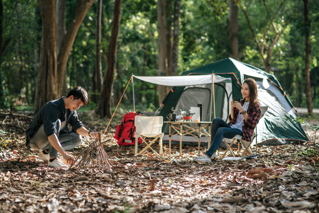 Young man picking branches and put them together, he preparing preparing a pile of firewood for fire camping on night and pretty girl friend sitting at front of camping tent